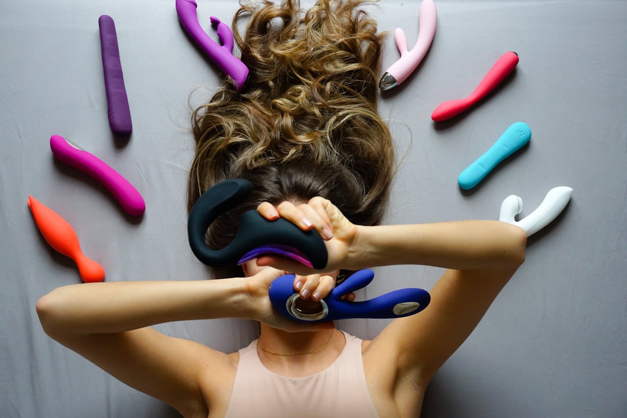 Woman lying on bed surrounded by colorful vibrators, expressing sensuality and pleasure.